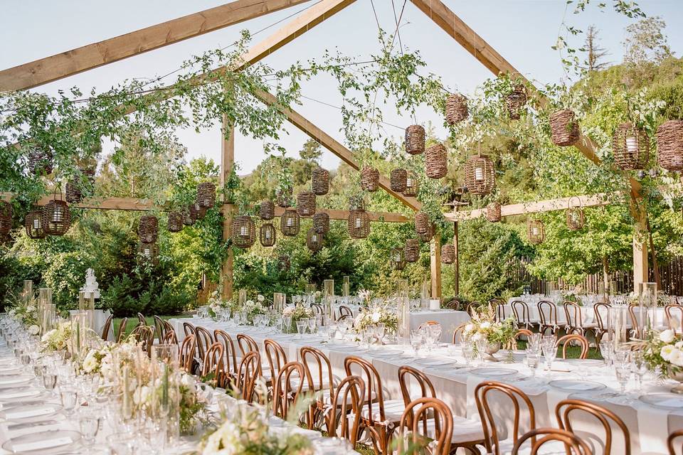 Large outdoor wedding reception dinner under a wooden pergola strung with rattan lanterns
