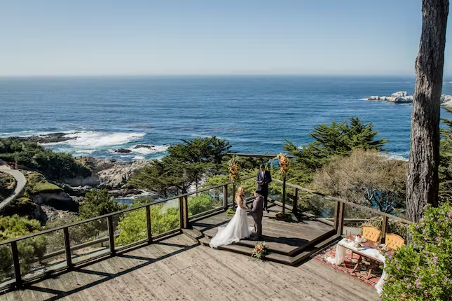 Bride in white dress and groom in black suit standing with officiant on a deck overlooking the breathtaking Big Sur coastline