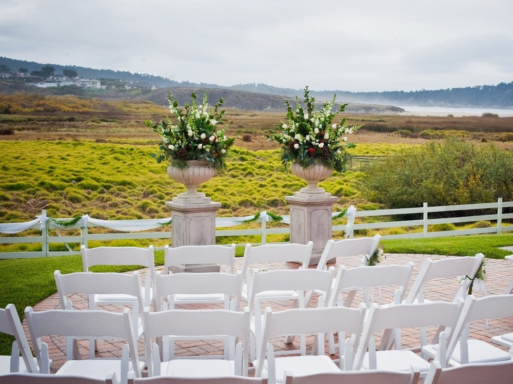 Large floral urn arrangements and white chairs set up on the Mission Ranch lawn with coastal views of Point Lobos in the distance