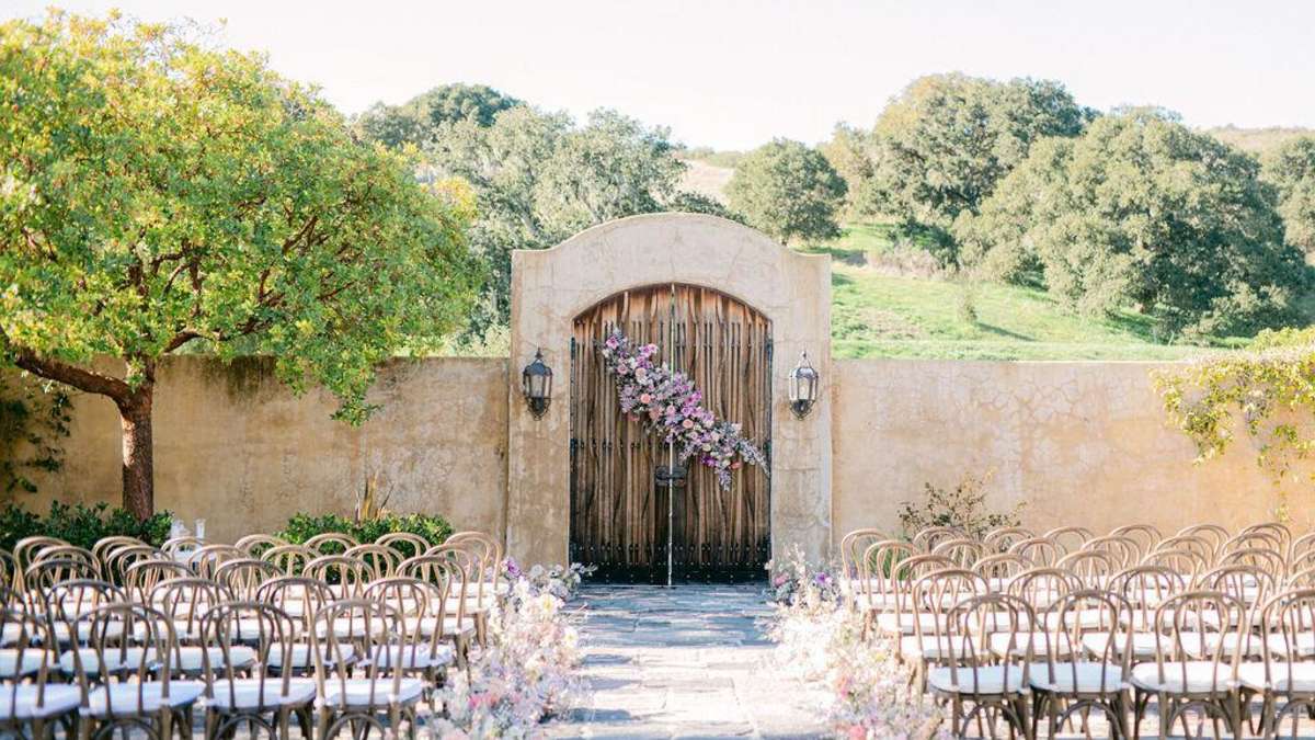 Outdoor wedding set up with mission-style adobe walls and large rustic wood gate covered with flowers