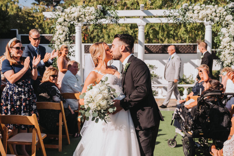 Bride and groom kissing after outdoor wedding ceremony in front of clapping audience and white arbor covered in flowers